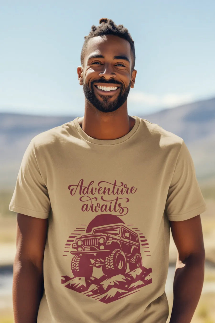 Man wearing a beige brown T-shirt with a rugged jeep and 'Adventure Awaits' text graphic, standing outdoors with a mountain background.