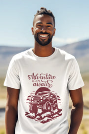 Man wearing a white T-shirt with a rugged jeep and 'Adventure Awaits' text graphic, standing outdoors with a mountain background.