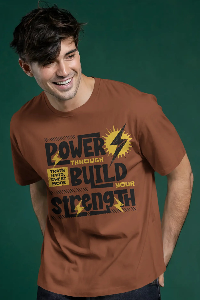 Man wearing a rust brown cotton t-shirt with “Power Through Build Your Strength” text graphic and yellow lightning bolts, smiling in front of green backdrop.